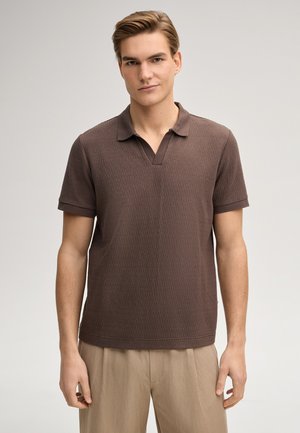 Junger Mann, trägt ein braunes, strukturiertes kurzärmeliges Polohemd und beige Hosen, steht vor einem schlichten hellen Hintergrund.