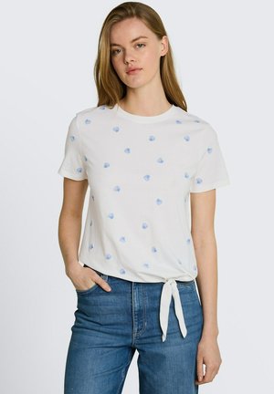 Femme portant une chemise blanche à manches courtes avec de petits imprimés coquillages bleus nouée à la taille, associée à un jean bleu, debout avec une main dans la poche.