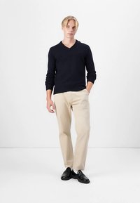 Marinblå v-ringad tröja i lättviktsstickning, kombinerad med beige raka byxor och svarta loafers; modellen står mot en enkel vit bakgrund.