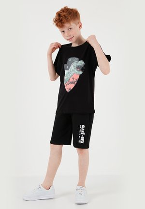 Chlapec s červenými vlasmi v čiernom tričku s témou dinosaurov a krátkych nohaviciach, ťahajúci golier, aby ukázal dizajn T-Rexa. Biele tenisky.