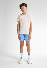 Camiseta de algodón crema con diseño gráfico azul, pantalones cortos azul claro, zapatos blancos y calcetines a rayas azul y blanco. Conjunto simple y casual.