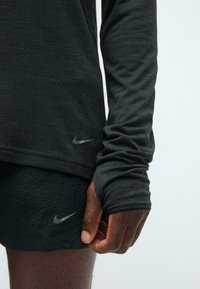 Crna dugorukava atletska majica s teksturiranim materijalom, sa suptilnim Nike logotipom. Osoba nosi usklađene crne kratke hlače, također s Nike logotipom.