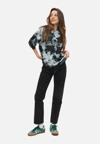 Černo-bílý tričko s motivem tie-dye a kruhovým logem, v kombinaci s černými džíny a zelenými teniskami s bílými pruhy. Model pózuje.