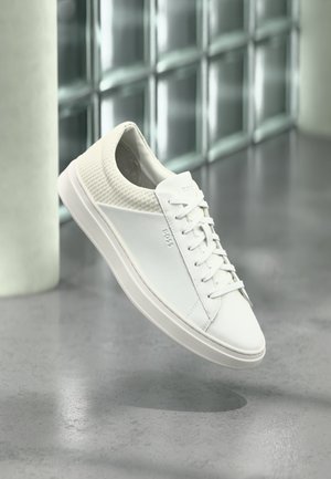 Weißer Low-Top-Sneaker mit gestepptem Fersenbereich und gebrandetem "BOSS"-Seitennlogo, dargestellt in der Luft schwebend auf grauem, poliertem Boden im Innenbereich.