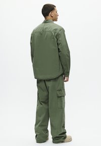 Completo cargo verde oliva con una camicia a maniche lunghe e pantaloni larghi, caratterizzato da numerose tasche e una silhouette rilassata.