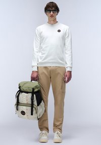 Sweatshirt en tricot blanc, pantalon cargo marron clair et un sac à dos multicolore avec des sections noires, vertes et beiges, et des détails en texte orange.
