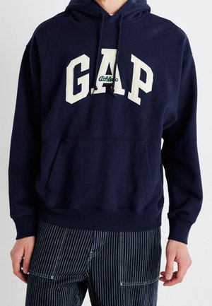 Mand iført marineblå hættetrøje med store hvide bogstaver "GAP" og lille grøn tekst "Athletic", kombineret med sorte bukser med tynde striber.