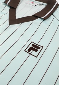 Chemise de sport bleu clair avec fines rayures verticales marron, col marron bordé d'une ligne blanche, et logo marron contouré sur la poitrine.