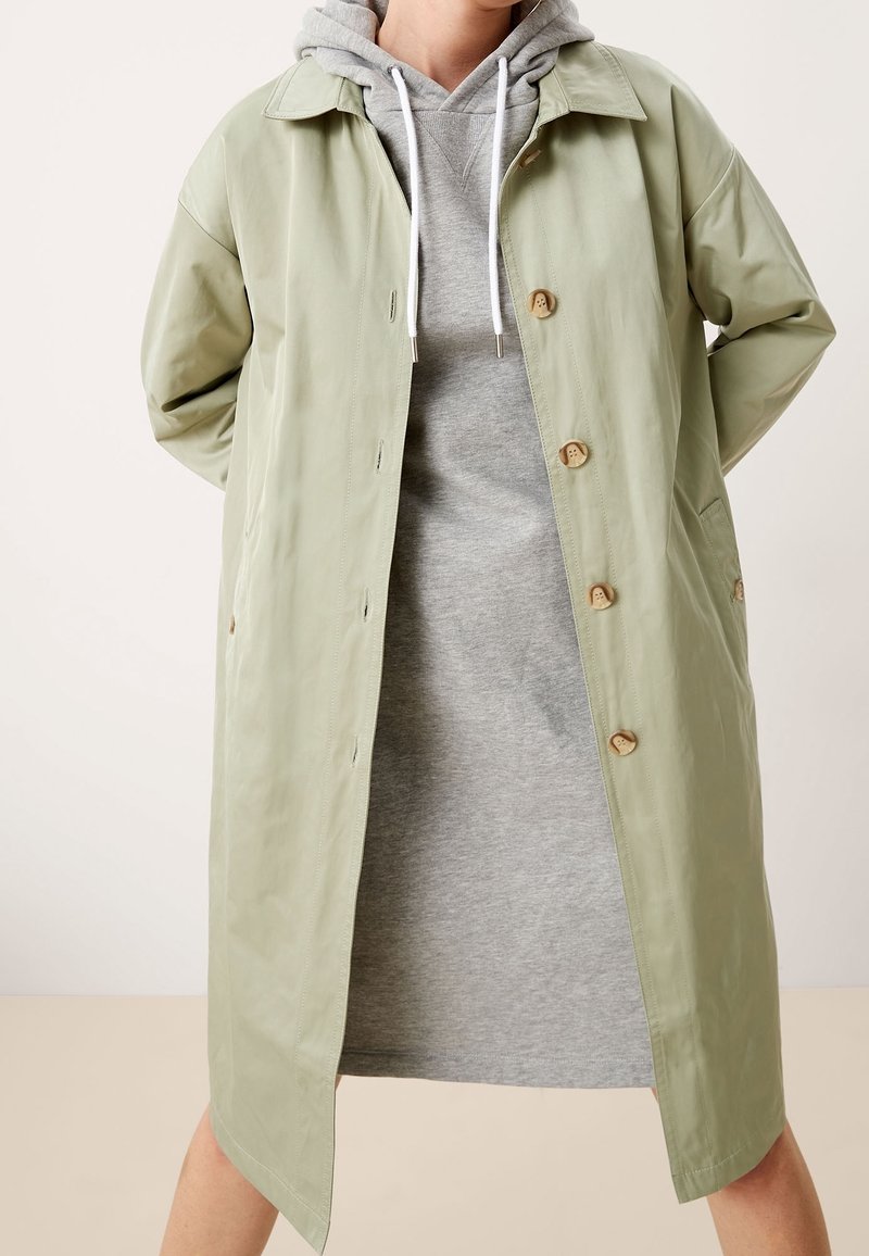 Personne portant un trench-coat vert clair boutonné sur une robe sweat à capuche grise, debout les mains dans le dos.