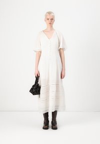 Robe blanche, longue jusqu'au sol, avec de courtes manches en dentelle, un devant à boutons et un design en couches ; portée avec un sac floral noir et des bottes à talons brunes.