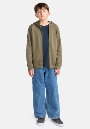 Haut à capuche olive zippé avec poches avant, porté sur un t-shirt bleu marine, associé à un jean baggy bleu clair et des baskets grises.