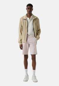 Veste beige clair, chemise blanche à boutons, shorts rose pastel, baskets blanches et chaussettes blanches. Design simple avec des motifs et textures minimaux.