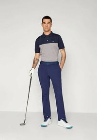 Námořnická a šedá polokošile s límečkem, spojená s námořnickými kalhotami. Model drží golfovou hůl, na nohou má bílé golfové boty s modrými akcenty.