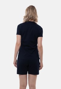 Person med axellångt blont hår som bär en enkel svart t-shirt med kort ärm och svarta shorts, vänd bort från kameran mot en vit bakgrund.