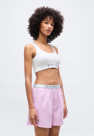 Γυναίκα με σγουρά μαλλιά που φοράει λευκό αθλητικό σουτιέν και ροζ ριγέ μποξέρ σορτς με το λογότυπο Polo Ralph Lauren.