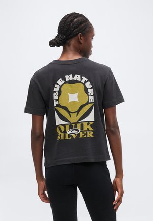 Άτομο με πλεξούδες που φοράει μαύρο T-shirt με ένα κίτρινο λουλούδι και το κείμενο "TRUE NATURE QUIKSILVER" στην πλάτη.