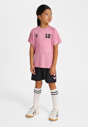 Ung pige iført pink sportsjakke med nummer 12, sorte shorts med chevron-design, knælange hvide sokker med sorte striber og hvide sneakers.