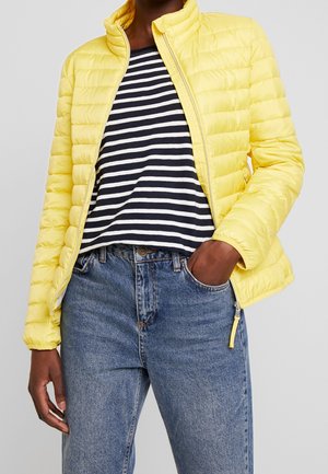 Personne portant une veste matelassée jaune sur un t-shirt rayé noir et blanc, un jean bleu, une main dans la poche.