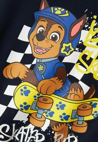 Personaggio di cane in stile cartone animato su uno sfondo scuro, indossa un casco blu e una divisa, mentre tiene uno skateboard giallo con impronte di zampe blu.
