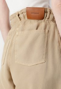 Beige bomullsbukser med elastisk midje, med to baklommer og en brun lærmerkel med preget tekst.