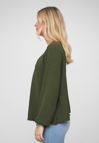 Olijfgroene long-sleeve top met een ontspannen pasvorm, textuurstof, ronde halslijn en licht uitlopende mouwen, gedragen met lichte spijkerbroek.