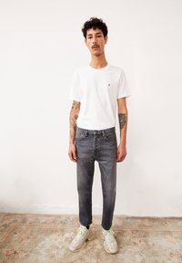 Weißes Baumwoll-T-Shirt mit kleinem lila Logo, kombiniert mit grauen Jeans und hellen Sneakers, steht auf einem gemusterten Teppich.