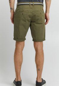 Shorts en coton vert olive avec des ourlets retroussés, des poches avant et arrière, et une taille contrastante. Portés avec des baskets noires.
