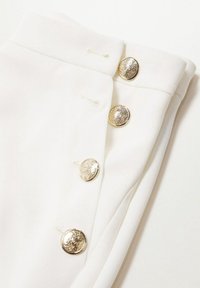 Witte stof met een gladde textuur, met vijf metallic gouden knopen langs de voornaad. De knopen hebben een ornamenteel ontwerp.