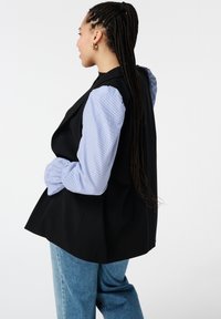 Zwarte mouwloze vest over een lichtblauw gestreept shirt met opgesmukte mouwen, gecombineerd met lichtblauwe denim. Minimalistisch ontwerp, gladde textuur.