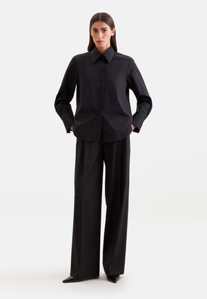 Vrouw staand met handen in de zakken, gekleed in een zwart overhemd met lange mouwen en wijde zwarte broek op een effen achtergrond.