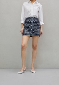 Camicia bianca a bottoni abbinata a una mini gonna blu navy con motivo geometrico e bottoni frontali, completata da tacchi bianchi.