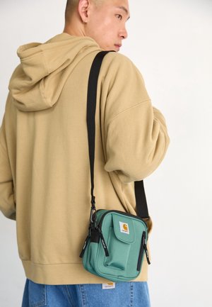 Homme en sweat à capuche beige et jean bleu portant un petit sac d'épaule vert avec une sangle noire, debout devant un fond blanc.