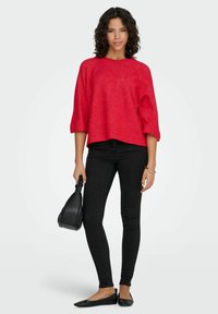 Ohlapen pulover rdeče teksture s tromesečnimi rokavi, kombiniran s črnimi oprijetimi kavbojkami in črnimi balerinkami, drži majhno črno torbico.