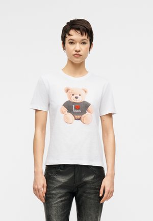 Donna con capelli corti e scuri che indossa una maglietta bianca con un orsetto che porta una maglietta nera con la scritta "I love Paris", abbinata a pantaloni neri.