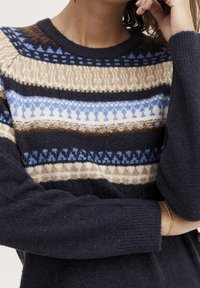 Femme portant un pull bleu marine avec des rayures à motifs beige, bleu et marron, main posée près du visage, bracelet en or visible au poignet.