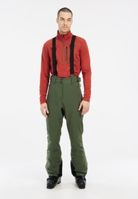 Chemise rouge à manches longues avec une poche zippée et pantalon vert imperméable avec des bretelles noires ajustables, présentant une texture lisse et une coupe ajustée.