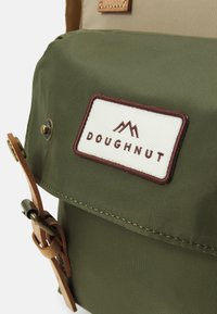 Green canvas backpack with brown leather accents and a white label reading "DOUGHNUT." Features a textured finish and brass hardware.