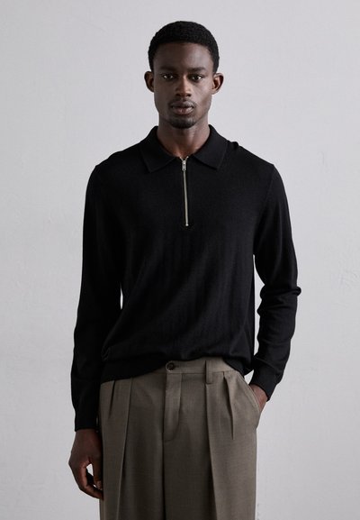 Sort strikket zip-necks pullover med krave-design. Teksturstof og lange ærmer, parret med beige plisserede bukser.