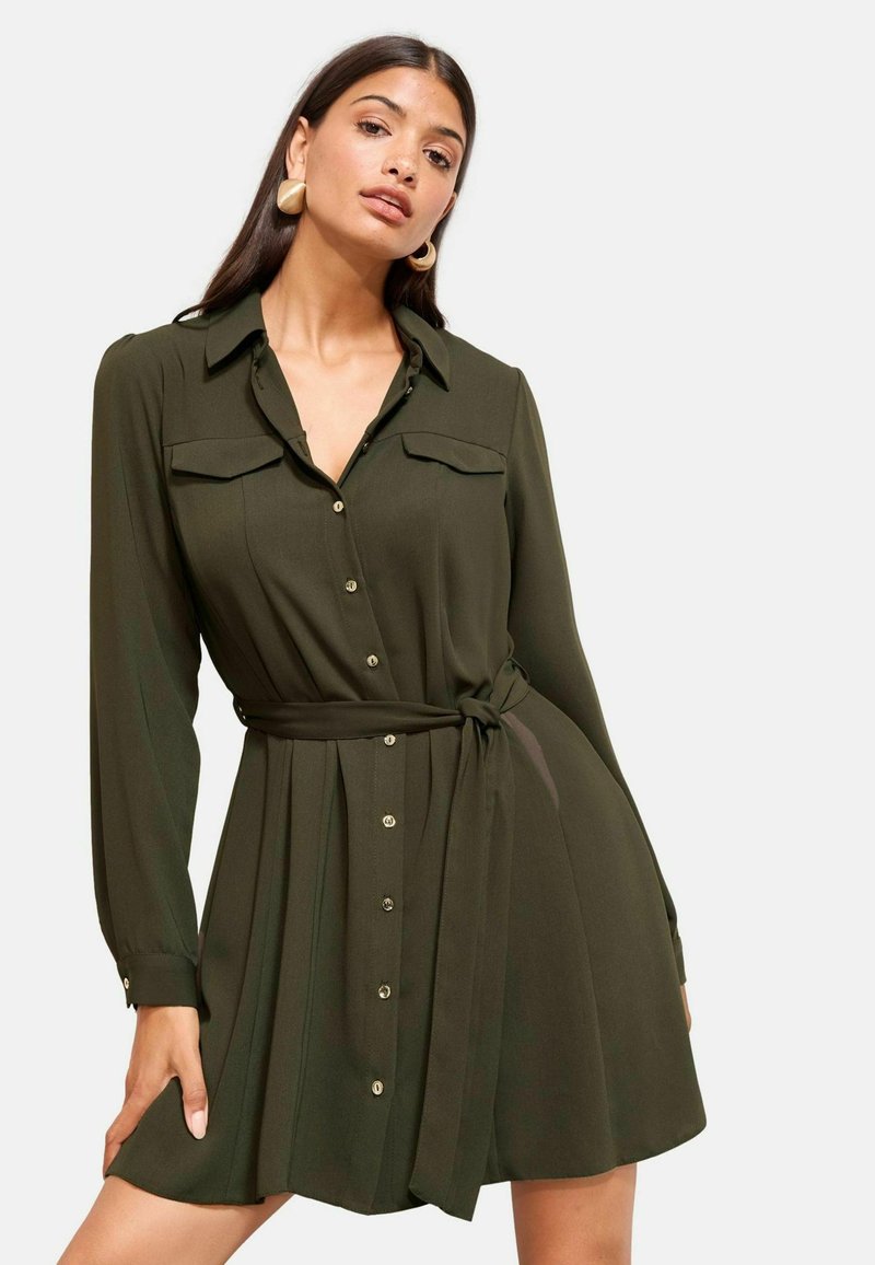 Rochie-cămașă verde măsliniu cu mâneci lungi, închidere cu nasturi în față, două buzunare la piept și talie cu curea. Prezintă o fustă fluidă și o textură netedă.