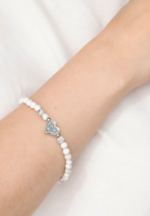 Wrist wearing a white beaded bracelet with a silver geometric heart charm, resting on a white fabric background.