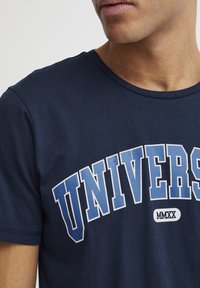 Tamsiai mėlyna medvilninė marškinėliai su baltais ir mėlynais "UNIVERS" užrašu su arkos dizainu, apvaliu kaklu ir trumpomis rankovėmis.