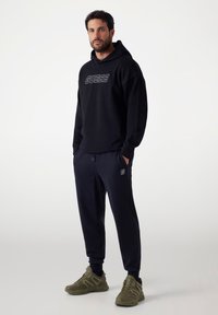 Čierna mikina s kapucňou a logom "GUESS", v kombinácii s tmavomodrými teplákmi so bočnými vreckami. Olivovo zelené tenisky. Jednoduchý, voľný strih.