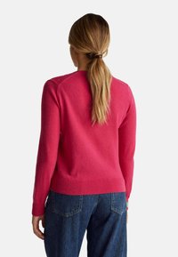 Donna con capelli lunghi raccolti, che indossa un maglione rosa acceso e jeans blu, vista di spalle su uno sfondo uniforme.