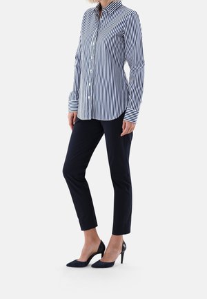 Femme portant une chemise boutonnée à manches longues rayée bleu et blanc, un pantalon noir ajusté et court, et des chaussures à talons hauts noires, debout sur un fond blanc.