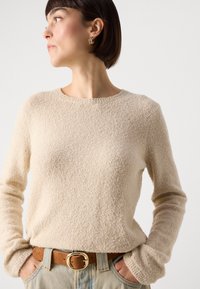 Beige textúrájú pulóver kerek nyakkivágással és hosszú ujjal, barna övvel és világoskék farmernadrággal párosítva.