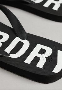 Des tongs noires avec une surface texturée, arborant des lettres blanches bien visibles. Les sangles sont noires et légèrement brillantes, avec des sections rembourrées.