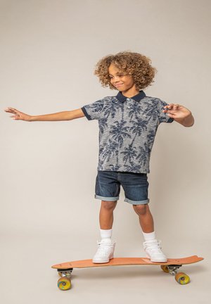 Enfant aux cheveux bouclés faisant de l'équilibre sur un skateboard orange, portant une chemise à motif de palmiers, un short en jean, des chaussettes blanches et des baskets.