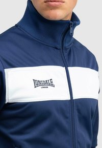 Nahaufnahme einer Person, die eine navyblaue Zip-Jacke mit einem hohen Kragen und einem weißen Brustpaneel mit dem Logo "Lonsdale London" trägt.