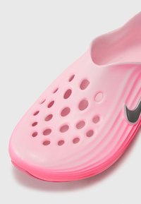 Rosa slip-on sko laget av lett materiale med perforert design. Har en glatt tekstur og en grå Nike-logo som aksent.