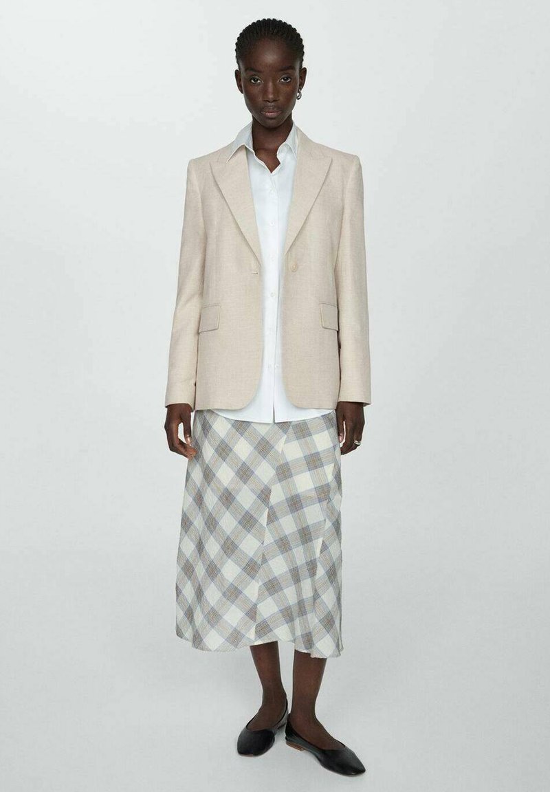 Beige Blazer mit scharfen Revers über einem weißen Hemd und einem karierten Wickelrock in Hellblau, Grau und Weiß. Schwarze flache Schuhe.